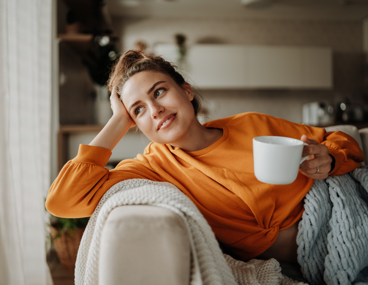 Eine junge Frau sitzt zugedeckt auf ihrem Sofa und sie hält eine Tasse in der Hand während sie aus dem Fenster schaut