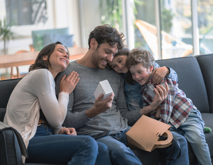 Eine Mama und ein Papa sitzen gemeinsam mit ihrem Sohn und ihrer Tochter auf dem Sofa übergeben Geschenke und umarmen sich