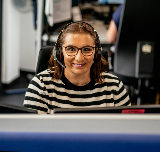 A woman with brown hair, wearing glasses and a black-and-white striped sweater, is smiling at the camera while seated at her desk in an office environment. She is wearing a headset. In the background, another person is working at a computer.