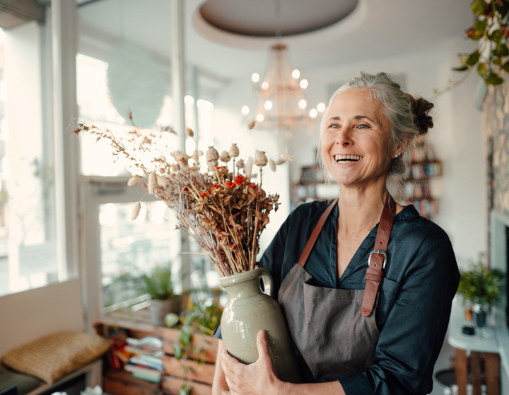 Een vrouw van middelbare leeftijd staat glimlachend in haar bloemenwinkel en houdt een vaas met droogbloemen in haar handen.