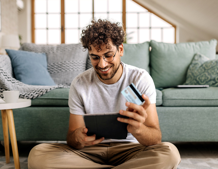 Een jonge man zit op zijn tablet in de woonkamer en houdt een creditcard in zijn hand