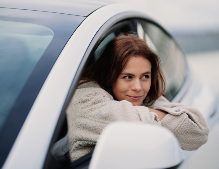 Eine junge Frau lehnt sich lächelt aus dem Fenster eines grauen Autos.